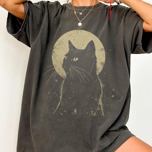 Puede incluir: Camiseta gris oscuro con un diseño de gato negro sobre un círculo marrón claro. El gato está representado de perfil, con bigotes. La camiseta tiene un aspecto vintage y desgastado, mangas cortas y un corte holgado. Ideal para uso casual.