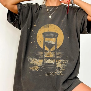 May include: Dark gray t-shirt featuring a large hourglass design with a yellow sun in the background. The hourglass is filled with sand, and the design has a vintage, distressed look. The shirt has short sleeves and a relaxed fit.