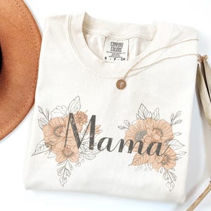 Puede incluir: Camiseta color crema con la palabra "Mama" en escritura gris oscuro, rodeada de ilustraciones florales melocotón y beige. También se ven un collar dorado con un colgante y un sombrero marrón.