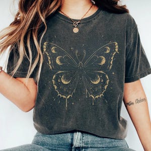 May include: Dark grey t-shirt featuring a gold butterfly design with moon phases on the wings. The butterfly is centered on the shirt, with a vintage, worn look. The shirt is paired with blue jeans.