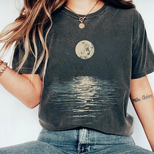 以下が含まれることがあります： 水面上の満月のグラフィックが特徴の濃いグレーのTシャツ。デザインには月、その反射、星空が含まれています。このシャツは半袖で、クルーネックです。