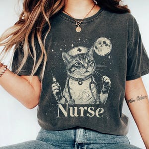 Puede incluir: Camiseta gris oscuro con un gato vestido de enfermera, con gorro, estetoscopio y jeringuilla. El diseño incluye una luna, estrellas y la palabra "Nurse" en una fuente vintage. Un diseño único y caprichoso.