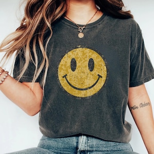 Puede incluir: Camiseta gris oscuro con un gran gráfico de cara sonriente amarilla desgastada. La camiseta tiene un aspecto vintage y desgastado y un cuello redondo clásico. La modelo lleva vaqueros azules y un collar.