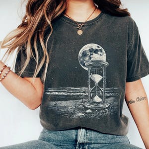 Puede incluir: Camiseta gris oscuro con un gráfico en blanco y negro de un reloj de arena, una luna llena y un paisaje marino. El diseño tiene una estética vintage. La camiseta tiene cuello redondo clásico y mangas cortas.