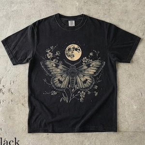 Op de afbeelding: Een zwart t-shirt met korte mouwen en een grafische print van een grote nachtvlinder onder een volle maan. De vlinder is omringd door delicate bloemillustraties. Het ontwerp is uitgevoerd in gedempte, lichte tinten op de donkere stof.