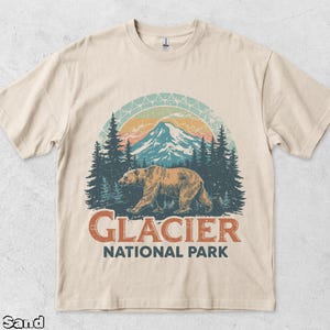 Puede incluir: Una camiseta de manga corta color arena con un gráfico de un oso pardo caminando frente a un bosque y una cordillera. El diseño incluye un arco de atardecer y el texto GLACIER NATIONAL PARK en letras de color naranja y azul oscuro.