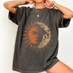 May include: A charcoal grey short-sleeve t-shirt featuring a large graphic print on the front. The design depicts a sun and moon combination, with one half showing a human face within a sun and the other half showing a textured crescent moon.