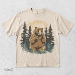 May include: A sand-coloured t-shirt featuring a graphic print of a brown bear playing a harmonica. The bear is centred in front of a large, pale yellow moon and a dark green forest of pine trees. The design is detailed with textured shading.