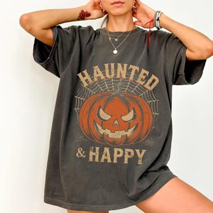 Puede incluir: Una camiseta gris carbón de manga corta con un gráfico de una calabaza naranja sobre un fondo de telaraña. El texto HAUNTED está impreso en forma de arco sobre la calabaza, y & HAPPY está impreso en una fuente recta debajo.