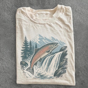 Puede incluir: Una camiseta de manga corta color crema con una ilustración gráfica de un salmón saltando sobre una cascada. La escena incluye montañas en tonos azules al fondo y pinos de color verde oscuro con agua salpicando alrededor.