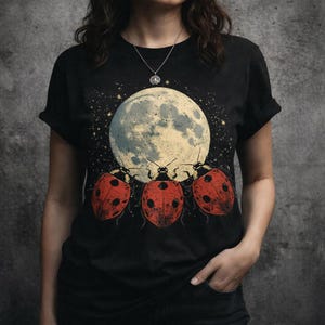 Puede incluir: Una camiseta negra de manga corta con un estampado gráfico de tres mariquitas rojas con puntos negros situadas bajo una gran luna llena de color amarillo pálido y textura, rodeada de pequeñas estrellas sobre un cielo nocturno oscuro.