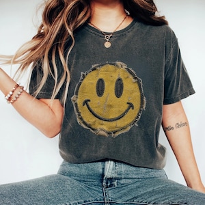 Puede incluir: Una camiseta de manga corta gris carbón con un gráfico grande de cara sonriente amarilla desgastada en el centro del pecho. El gráfico tiene una apariencia texturizada con bordes deshilachados y contornos bordados en gris oscuro para los ojos y la boca.