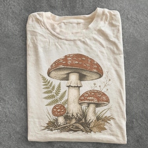 Op de afbeelding: Een crèmekleurig T-shirt met korte mouwen en een gedetailleerde, aardekleurige grafische print van drie paddenstoelen van verschillende groottes, vergezeld door een varenblad, dunne grassprieten en een gevallen blad aan de voet van de illustratie.