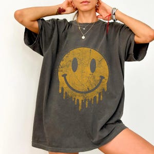 Puede incluir: Una camiseta de manga corta gris carbón con un gráfico grande de una cara sonriente amarilla desgastada en la parte delantera. El diseño circular tiene una textura agrietada y parece derretirse en el borde inferior, creando un efecto de goteo.