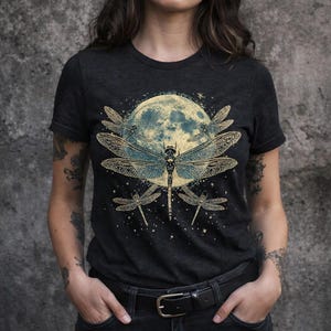 Puede incluir: Una camiseta de manga corta gris carbón con un estampado gráfico de una gran libélula central con alas detalladas, rodeada por tres libélulas más pequeñas y una luna amarilla grande y texturizada sobre un cielo nocturno estrellado.
