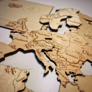 Könnte beinhalten: Eine Holzkarte von Europa mit eingravierten Ländernamen. Die Karte besteht aus mehreren Teilen, wobei Länder wie Frankreich, Deutschland und Italien deutlich sichtbar sind. Die Karte hat eine hellbraune Farbe.