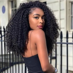 May include: A woman with dark skin and long, curly black hair. She is wearing a black spaghetti strap top and looking over her shoulder. A silver watch is on her wrist. The background includes a black fence and a white building.