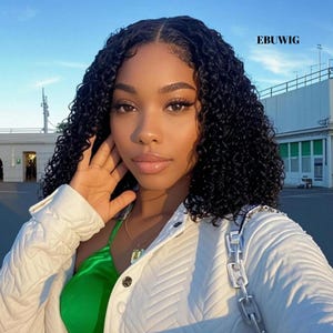 May include: A person with dark skin and curly black hair. They are wearing a white quilted jacket and a green top. The image is taken outdoors with a blue sky in the background. The text "EBUWIG" is visible in the upper right corner.