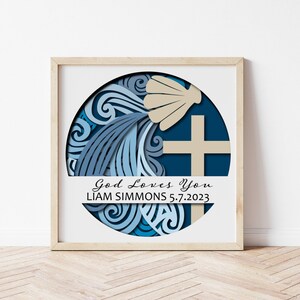 Può includere: Un'opera d'arte incorniciata con un design a strati. Un design circolare blu e bianco presenta una croce, acqua e una conchiglia. Il testo "God Loves You" e un nome e una data sono stampati su uno striscione bianco.
