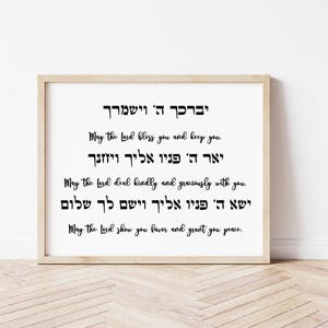 May include: A framed print with Hebrew and English text. The text is in black against a white background. The frame is a light wood color. The text includes phrases like "May the Lord bless you and keep you."