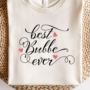 May include: Cream-colored t-shirt with the phrase "best Bubbe ever" in elegant black script. Small pink hearts accent the text. The shirt is folded on a woven surface.