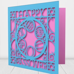 May include: A blue laser-cut greeting card with the words "HAPPY PASSOVER" and a central circular design with symbolic elements. The card has a pink interior and is displayed at an angle.