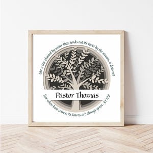 May include: A framed art print featuring a layered paper tree design in shades of gray and white. The text "Pastor Thomas" is centered below the tree, with a quote encircling the tree. The frame is a light wood color.