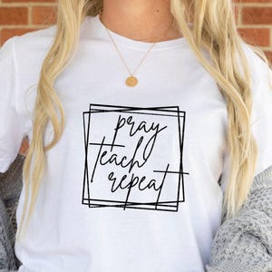 Op de afbeelding: Wit T-shirt met de woorden "pray teach repeat" in een zwarte, handgeschreven lettertype in een zwart vierkant. Het shirt heeft een ronde hals.