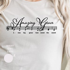 May include: Cream-colored long-sleeve shirt featuring the text "Amazing Grace" in a black, elegant script above a musical staff. The staff includes musical notes and the words "A - maz ing grace how sweet the sound". The shirt also has the text "NIGHT OWL LANE".