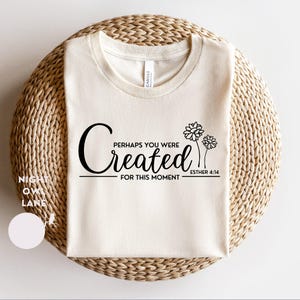 May include: Cream-colored t-shirt with the text "Perhaps You Were Created For This Moment" and Esther 4:14. The design includes stylized flowers. The shirt is displayed on a woven, circular surface.