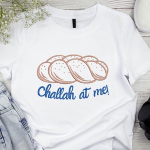 Puede incluir: Camiseta blanca con un diseño bordado de pan challah marrón, con pequeños puntos. El texto "Challah at me!" está escrito en azul debajo. La camiseta es de estilo casual.