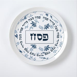 May include: White ceramic plate with a blue floral and text design. The plate features a central rectangular text box with Hebrew characters, surrounded by a circular pattern of leaves and additional Hebrew text.