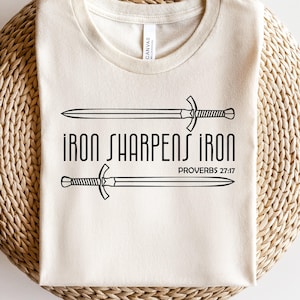 Pode incluir: Camiseta creme com o texto preto "IRON SHARPENS IRON" e "PROVÉRBIOS 27:17". Duas espadas estão acima e abaixo do texto. A camiseta é feita de material macio e é adequada para uso casual.