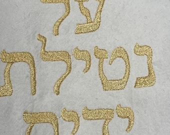 May include: Gold embroidered Hebrew text on a white fabric background. The text is in a stylized font, with each letter meticulously stitched. The embroidery thread has a metallic sheen, creating a contrast against the white fabric.