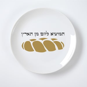 May include: White ceramic plate featuring a gold-coloured challah bread design and Hebrew text. The plate has a simple, elegant design, suitable for serving or display. The text is in black and is centred above the bread illustration.