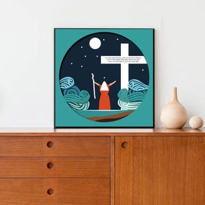 May include: Framed art print with a paper cut design. It depicts a figure in an orange robe and white head covering, arms raised towards a white cross. The background is a dark blue night sky with a full moon and stars. Text is visible on the cross.