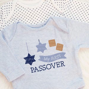 May include: Two baby onesies. The front onesie is light blue with a navy blue star and the words "My First Passover". The design includes two matzah crackers and a banner. The second onesie is white with a pattern of small navy blue stars.