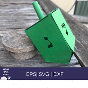 May include: A green dreidel with black Hebrew letters and symbols. The dreidel is made of folded paper and has a green handle. The image also includes a small, round, grey stone object and the text "EPS | SVG | DXF".