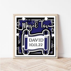 May include: A framed, layered paper art piece with a navy blue background. The design features the words "Mazel Tov" in white script above a scroll with the name "David" and the date "10.11.22". White stars surround the design.