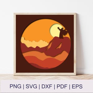May include: Framed paper art showing a layered landscape with a figure on a mountain, a sun, and clouds. The colour palette includes shades of orange, brown, and yellow. The bottom of the frame includes the text: PNG | SVG | DXF | PDF | EPS.