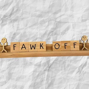 May include: Wooden block letters spell out "FAWK OFF" on a small wooden shelf, flanked by two small wooden figures. The letters are in a sans-serif font. The background is a textured, off-white surface.
