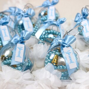 May include: Clear plastic ornaments filled with light blue beads and decorative elements. Each ornament is tied with a light blue ribbon and a tag that says "Yousuf". The ornaments are resting on a bed of white fabric flowers.