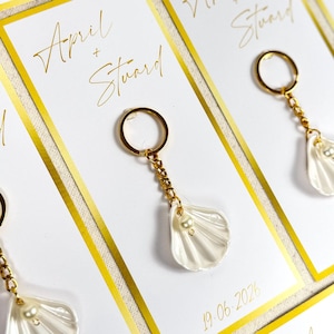 Może przedstawiać: Zbliżenie na upominki ślubne. Każdy upominek to zawieszka w kształcie muszli z perłą, przymocowana do złotego breloka. Upominki są prezentowane na białych kartach ze złotymi obramowaniami, imionami "April + Stuart" i datą "19.06.2026" w kolorze złotym.