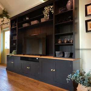 May include: A dark grey entertainment unit with a light wood top and brass handles. It has a TV, open shelves, and cupboards. The shelves display plants and baskets, offering a stylish storage solution. The unit is a modern take on a classic design.