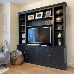 May include: A dark grey entertainment unit with a TV, shelves, and cupboards. The shelves hold framed pictures, ornaments, and a clock. A grey sofa and a wicker basket are in the foreground.