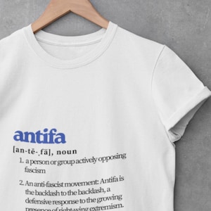 Può includere: T-shirt bianca con la parola "antifa" in blu, seguita da una definizione. La definizione include la frase "una persona o un gruppo che si oppone attivamente al fascismo" e una citazione di Todd Gitlin.