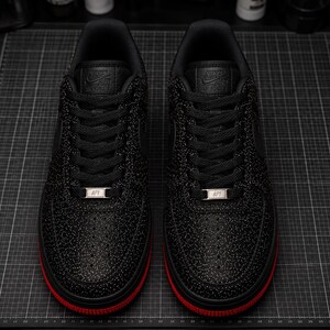 May include: Pair of black sneakers with a textured pattern and red soles. The sneakers have black laces and a silver metal tag that reads "AF1". The tongue of the shoe has the Nike Air Force logo.