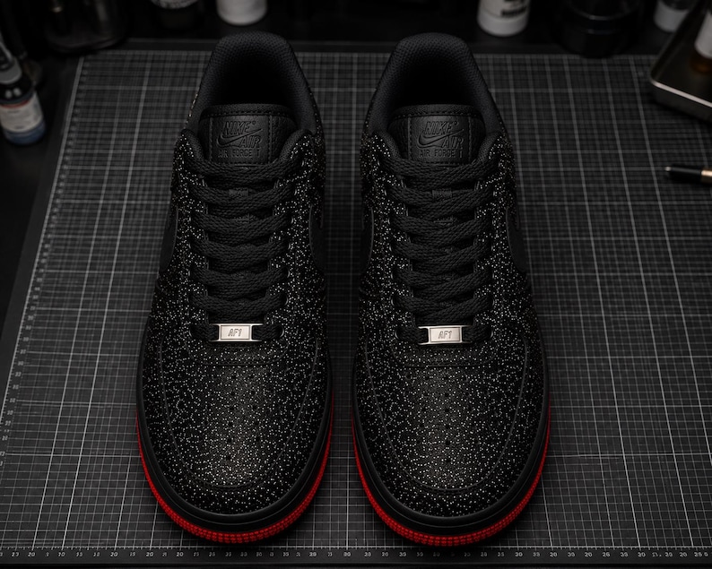 May include: Pair of black sneakers with a textured pattern and red soles. The sneakers have black laces and a silver metal tag that reads "AF1". The tongue of the shoe has the Nike Air Force logo.