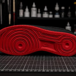 May include: Black and red Nike Air Force 1 sneaker sole. The sole features a vibrant red color with concentric circles and a textured pattern. The Nike logo is visible. The shoe is on a black grid surface.