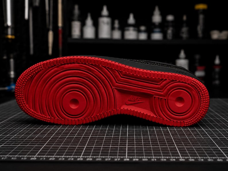 May include: Black and red Nike Air Force 1 sneaker sole. The sole features a vibrant red color with concentric circles and a textured pattern. The Nike logo is visible. The shoe is on a black grid surface.